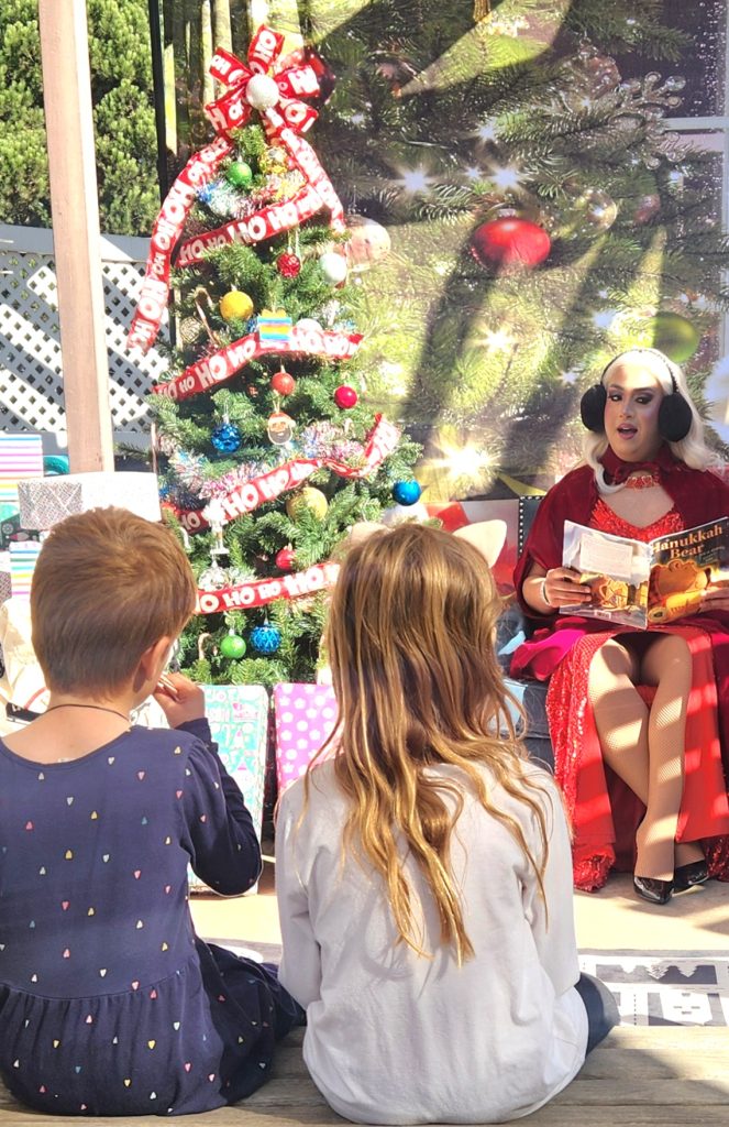 Mrs. Claus reading from santa's seat by the tree to 2 kids facing her (not the camera)